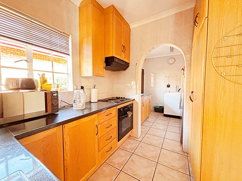 A functional kitchen featuring wooden cabinetry, built-in oven and hob, tiled splashbacks, and an arched transition to a secondary utility or laundry area.
