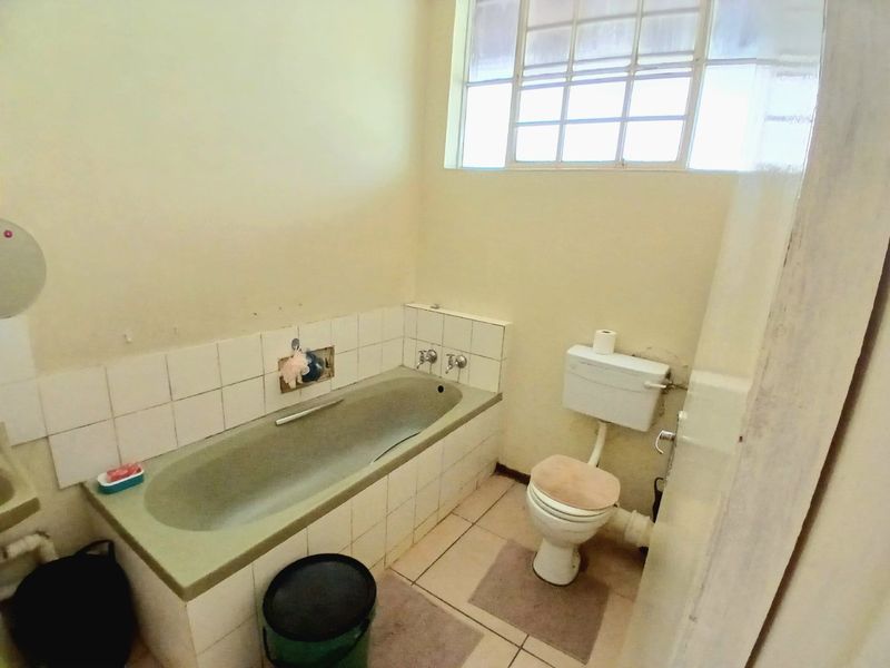 A dated bathroom featuring a built-in bathtub with tiled splashback, a standard toilet, and a high-set window providing natural light.