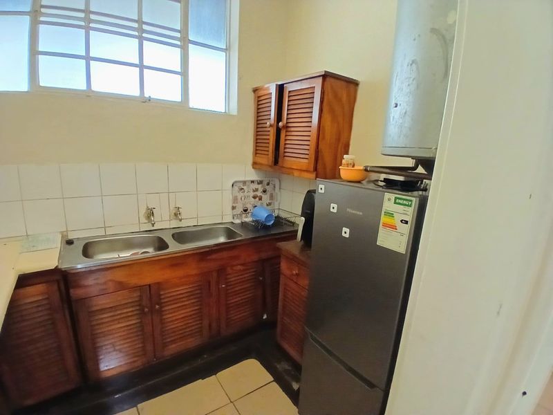 A compact kitchen featuring wooden cabinetry, a double stainless steel sink, tiled backsplash, and a wall-mounted geyser.