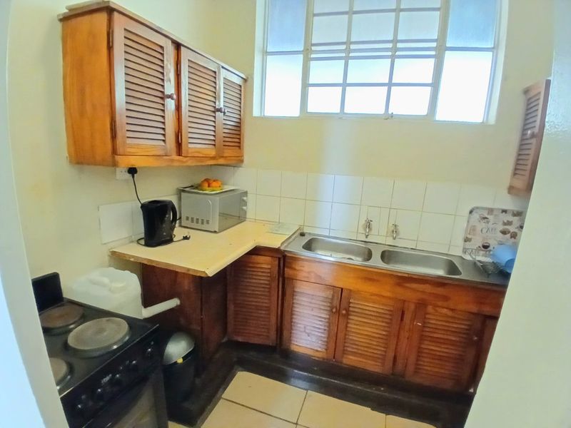 A compact kitchen featuring dated louvered timber cabinetry, a double bowl stainless steel sink, tiled splashback, and ceramic floor tiles.
