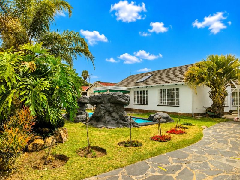 A well-maintained backyard featuring a swimming pool with decorative rock features, a stone-paved pathway, and established garden landscaping.