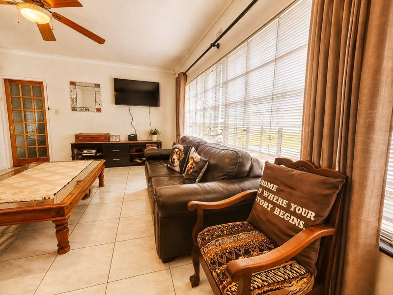 A comfortable living room featuring tiled flooring, a ceiling fan, and large windows with venetian blinds and curtains, providing ample natural light.