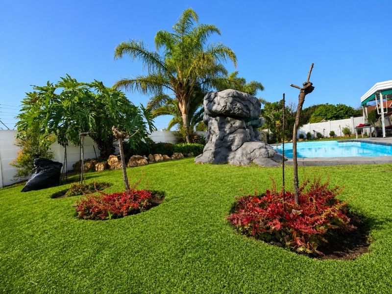 A well-maintained backyard featuring a swimming pool, a decorative artificial rock feature, and landscaped garden beds with established palm trees.