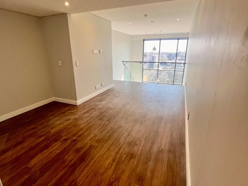 An empty, modern living area featuring warm-toned laminate wood flooring, recessed lighting, and a large window with a glass balustrade overlooking the city.