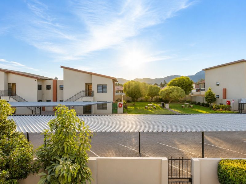 A view of the apartment complex exterior featuring a paved parking area with carports, a communal garden, and mountain views in the background.