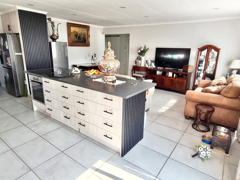 Modern open-plan living area featuring a central kitchen island with built-in oven, hob, and contemporary vertical slat detailing, complemented by neutral tiled flooring.
