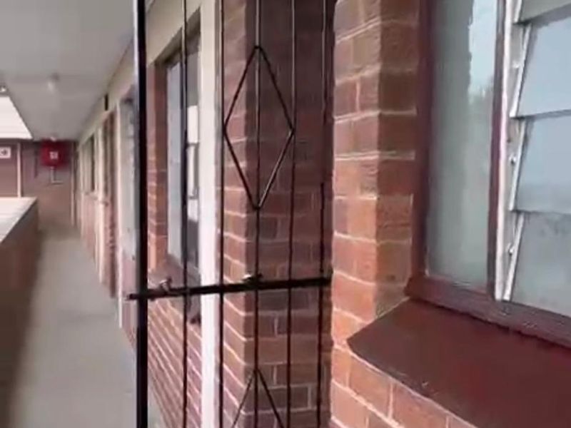 Exterior apartment hallway featuring brick facade, security gate, and louvre windows along a concrete walkway.