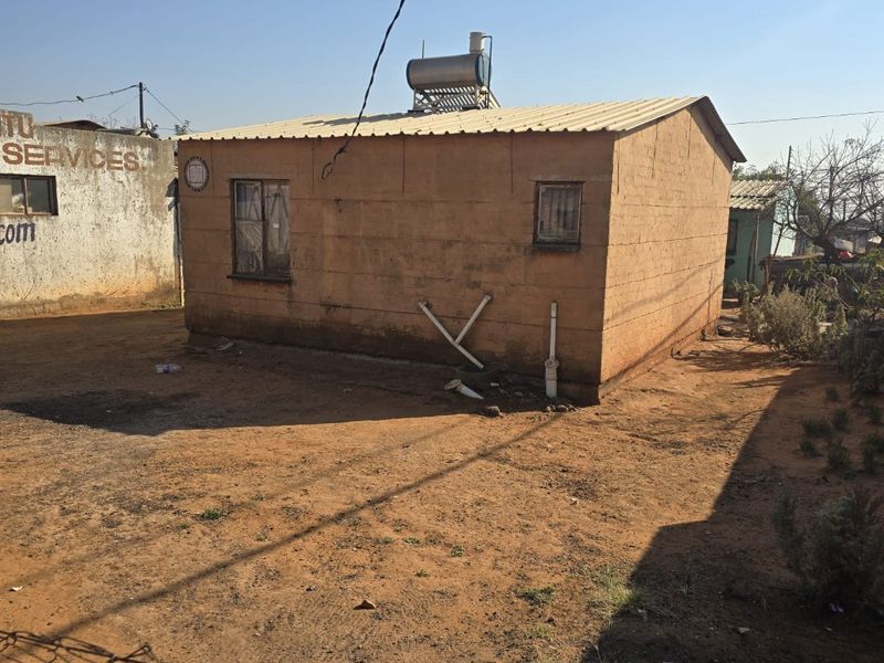 Rear view of a single-story dwelling featuring a corrugated metal roof, solar water heater, and exposed exterior plumbing on a dirt lot.