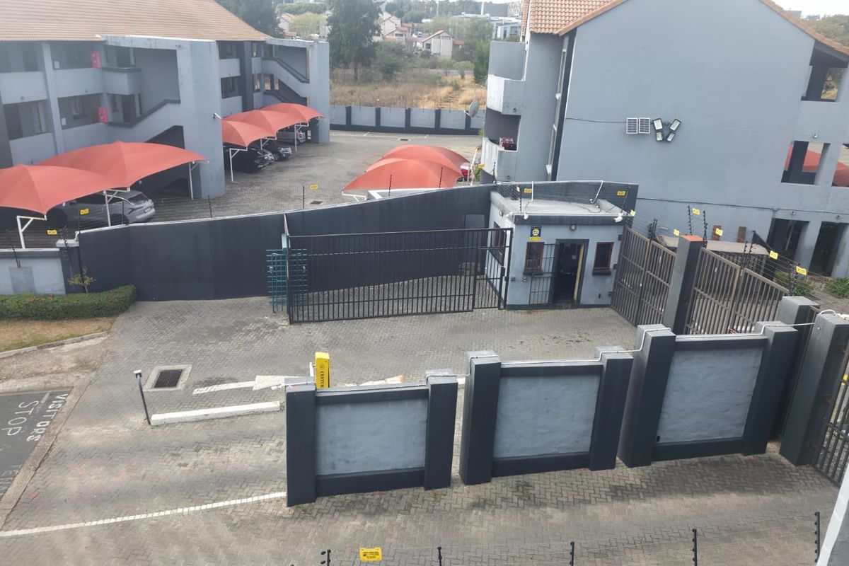 Elevated view of the apartment complex entrance featuring a security gate, guard house, paved driveway, and covered parking bays.