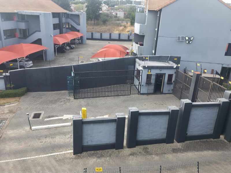 Elevated view of the apartment complex entrance featuring a security gate, guard house, paved driveway, and covered parking bays.