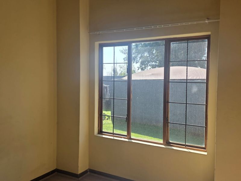 An empty bedroom featuring a large window with security bars and a curtain rail, providing natural light and a view of the exterior.
