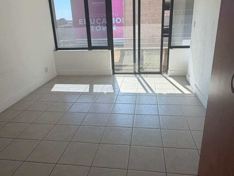 An empty, sunlit living area featuring neutral tiled flooring, recessed ceiling lights, and a sliding glass door leading to an exterior balcony.