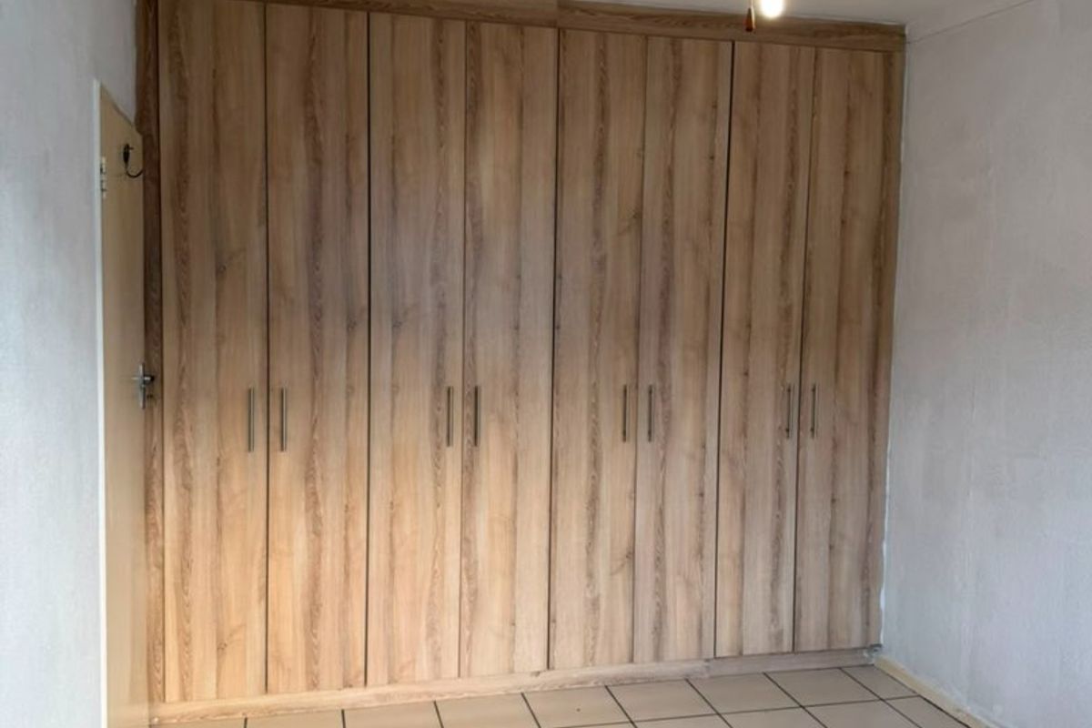 An empty bedroom featuring extensive built-in wooden cupboards, a ceiling fan, and tiled flooring, offering a blank canvas for personalization.