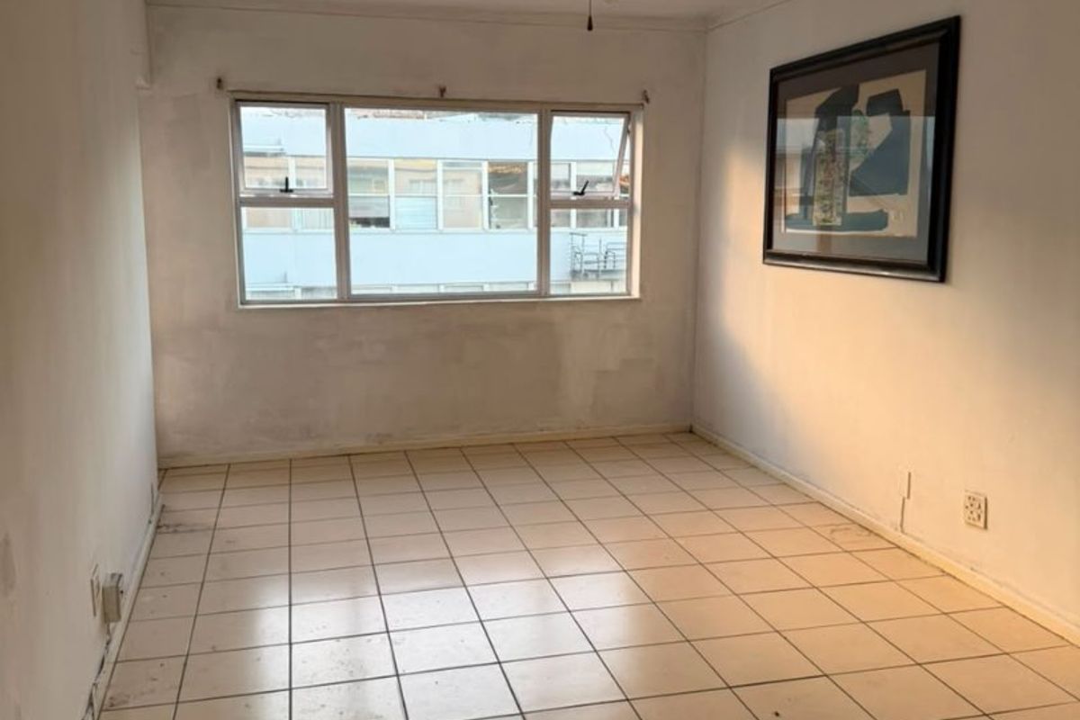 An empty living room featuring tiled flooring, a ceiling fan, and large windows, requiring cosmetic updates to the walls and flooring.