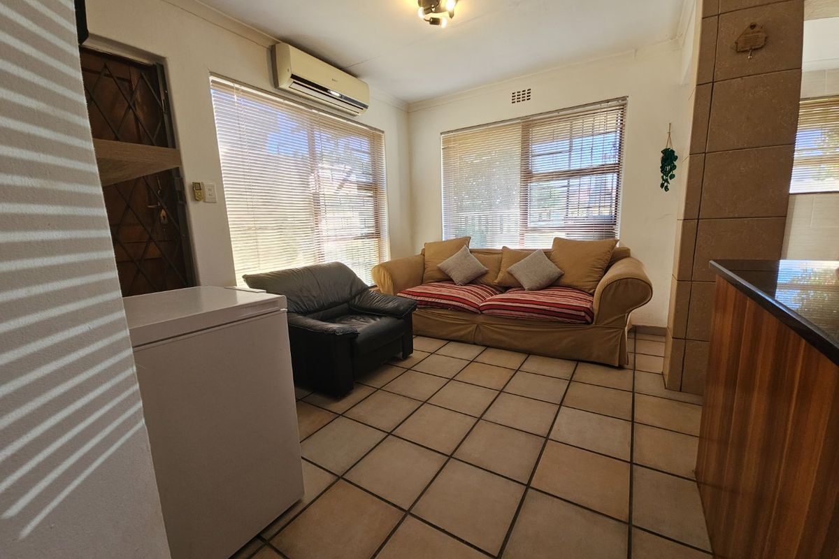 A functional living area featuring tiled flooring, a wall-mounted air conditioning unit, and large windows with venetian blinds.