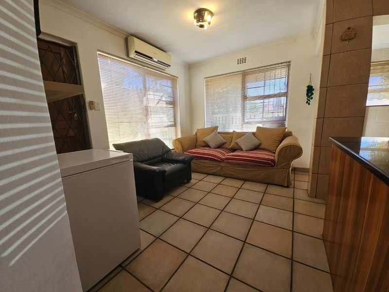 A functional living area featuring tiled flooring, a wall-mounted air conditioning unit, and large windows with venetian blinds.