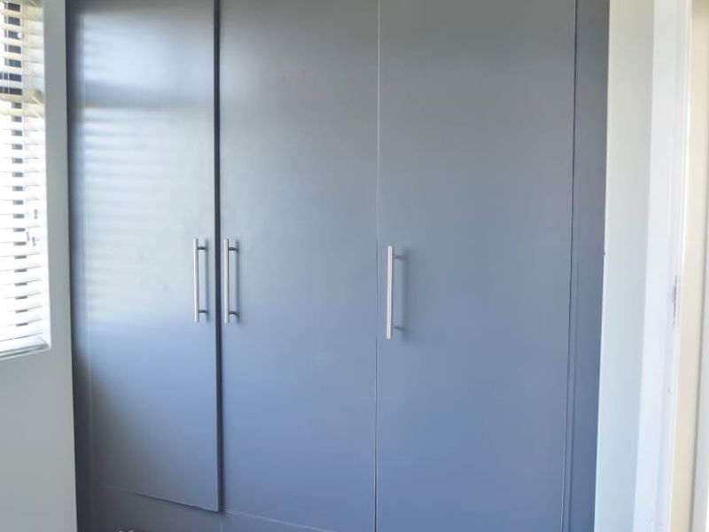 A clean, modern bedroom featuring floor-to-ceiling built-in cupboards with a matte grey finish, tiled flooring, and venetian blinds.