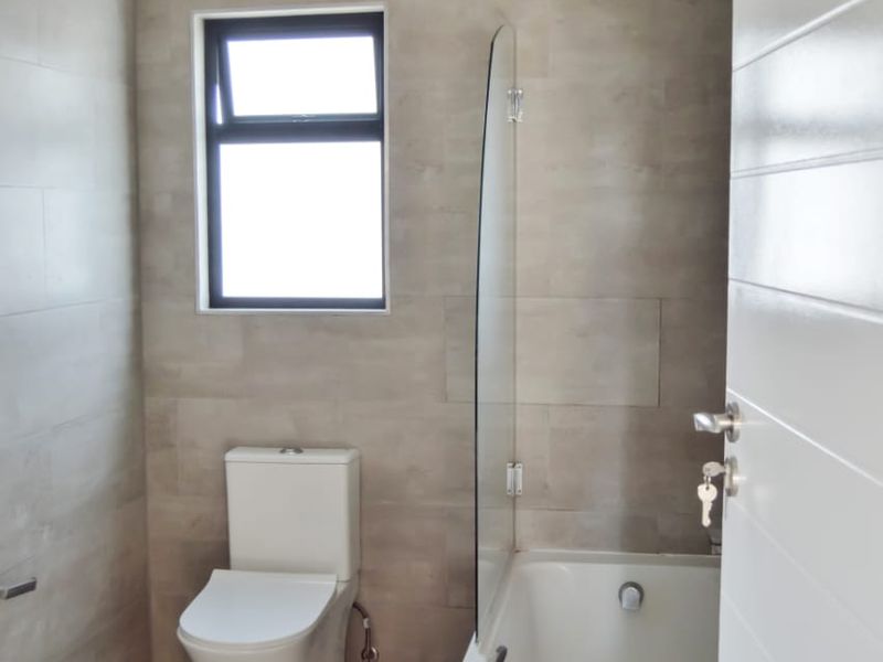 A modern, neutral-toned bathroom featuring a bathtub with a glass screen, a wall-mounted toilet, and floor-to-ceiling tiling.