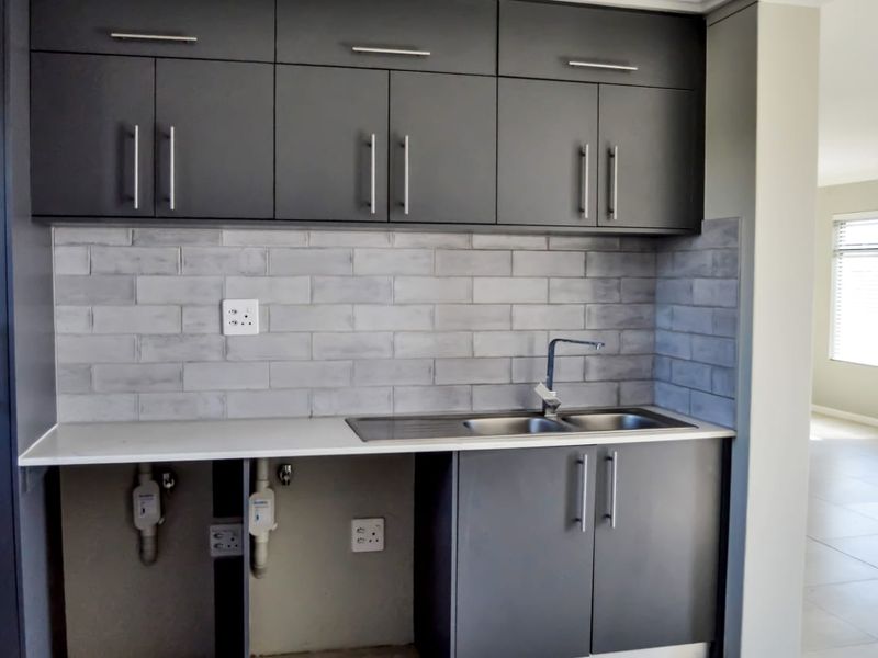 A functional scullery area featuring charcoal cabinetry, a subway tile backsplash, and a stainless steel double sink with plumbing connections for appliances.