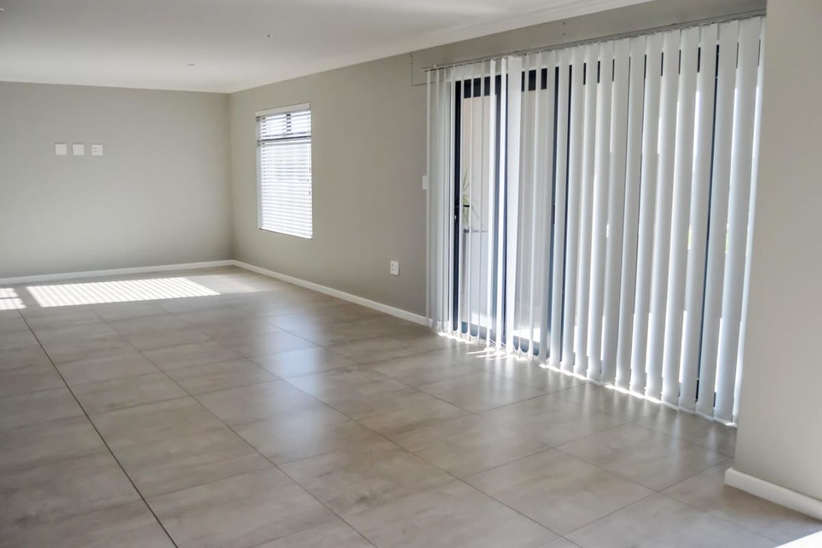 A bright, empty living room featuring neutral tiled flooring, vertical blinds, and a sliding glass door providing access to the exterior.