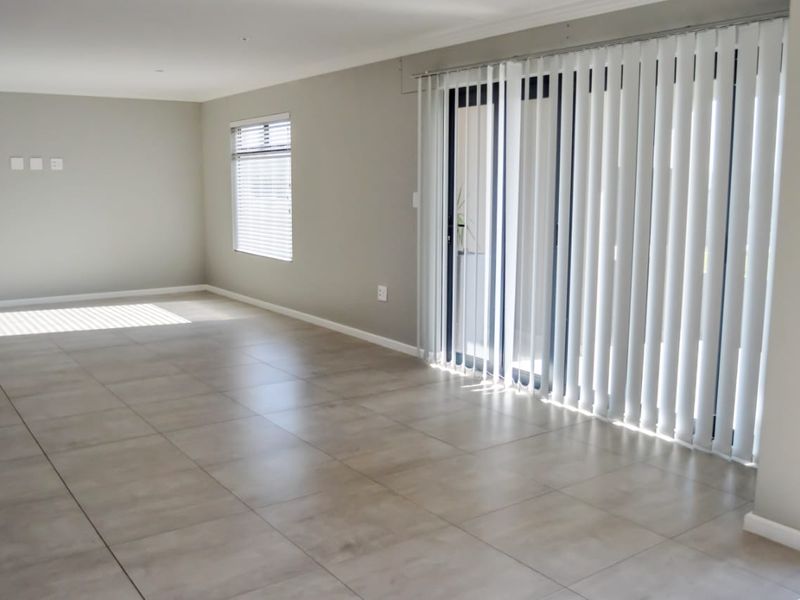 A bright, empty living room featuring neutral tiled flooring, vertical blinds, and a sliding glass door providing access to the exterior.