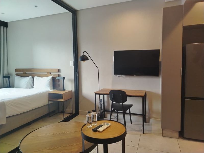 Modern open-plan apartment interior featuring a bedroom area separated by a sleek glass partition, tiled floors, and neutral finishes.