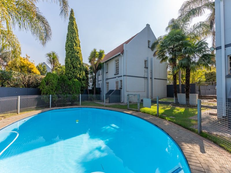 A well-maintained swimming pool area featuring a paved surround, safety fencing, and established garden landscaping set against the apartment building exterior.