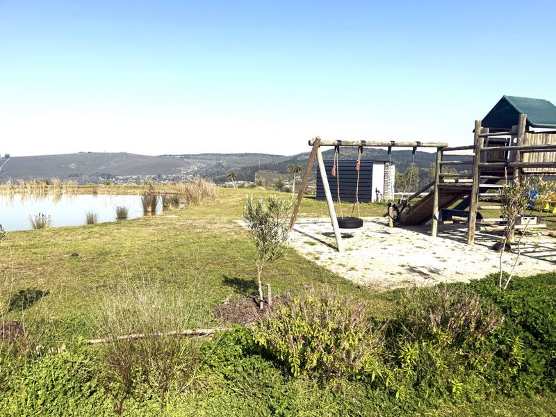 A spacious outdoor garden area featuring a natural pond, a wooden playground structure, and a small storage shed with scenic landscape views.