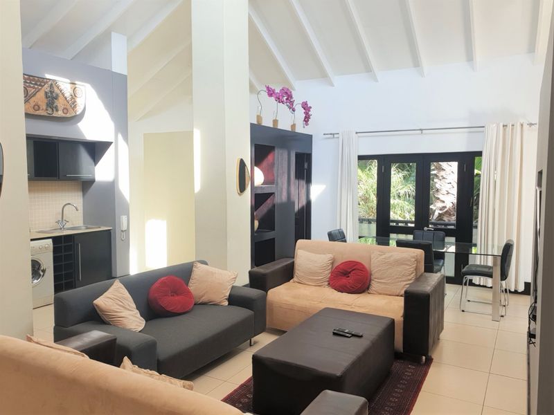 Spacious open-plan living area featuring vaulted ceilings with exposed beams, tiled flooring, and a compact integrated kitchen with direct access to outdoor space.