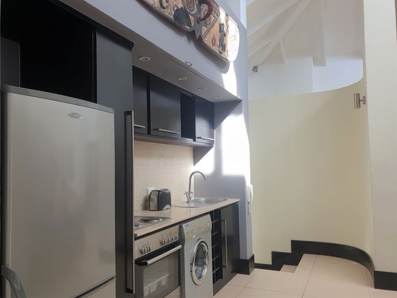 Compact kitchen area featuring dark cabinetry, integrated appliances, stainless steel sink, and tiled flooring, set within an open-plan living space with high ceilings.