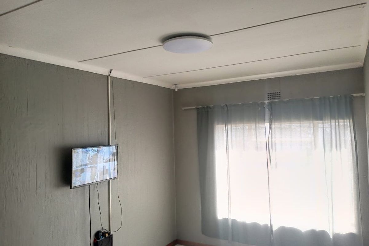 A sparsely furnished living area featuring tiled flooring, a ceiling light fixture, and neutral grey walls with surface-mounted electrical wiring.