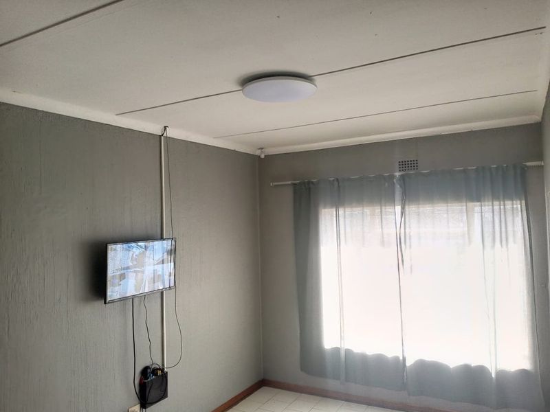 A sparsely furnished living area featuring tiled flooring, a ceiling light fixture, and neutral grey walls with surface-mounted electrical wiring.
