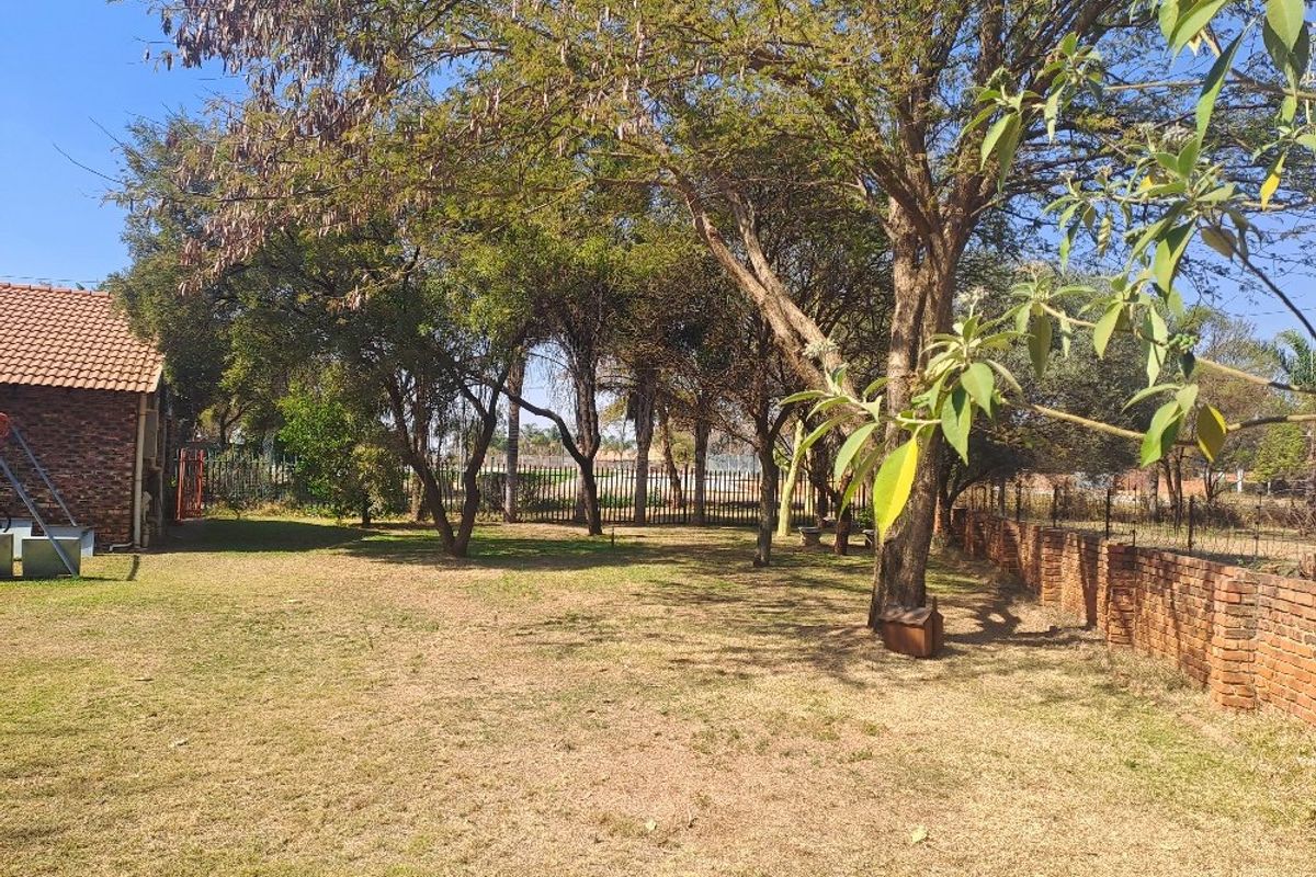 A spacious garden area featuring mature trees, a brick boundary wall, and metal perimeter fencing, adjacent to a building with a tiled roof.