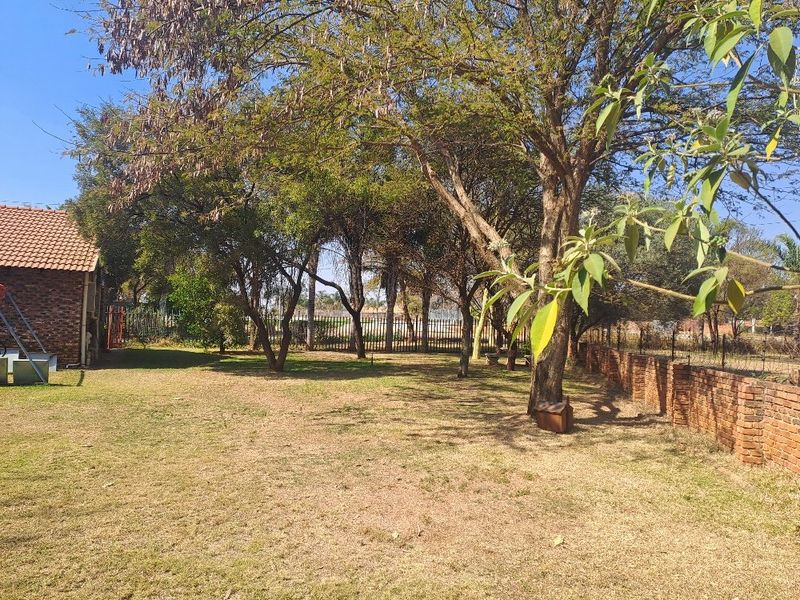 A spacious garden area featuring mature trees, a brick boundary wall, and metal perimeter fencing, adjacent to a building with a tiled roof.