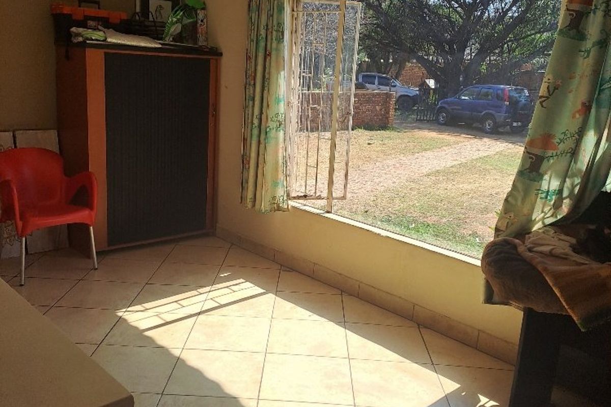 A living area featuring tiled flooring and a large window with security bars, offering natural light and views of the property exterior.
