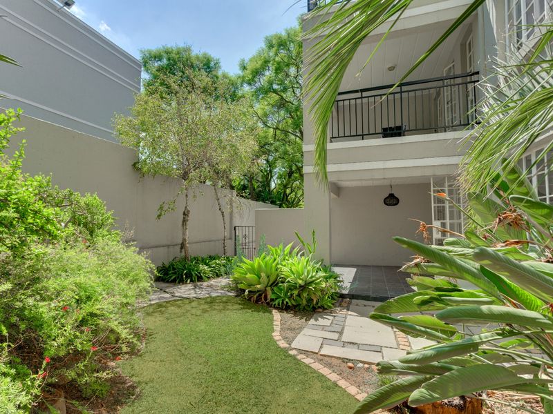 A private rear garden featuring a paved walkway, landscaped greenery, and a covered patio area with an overhead balcony.