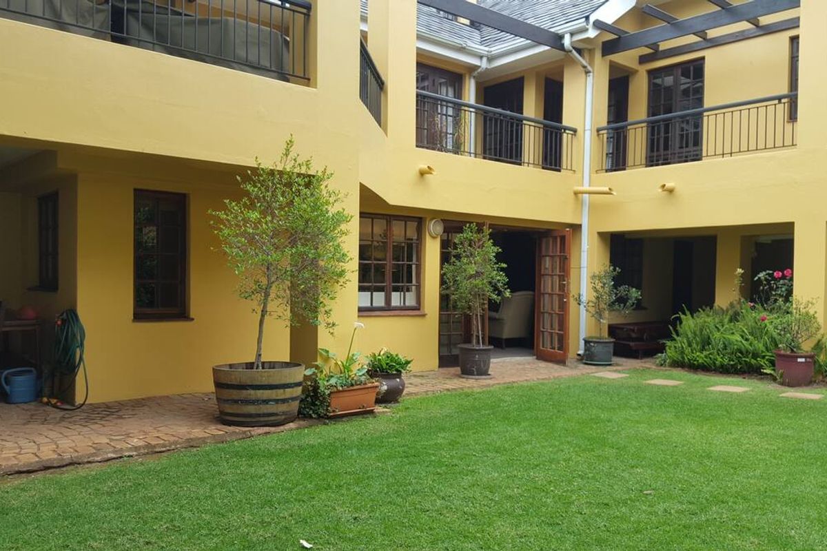 A rear courtyard featuring a manicured lawn, paved patio area, and a two-story building facade with balconies and timber pergola structures.