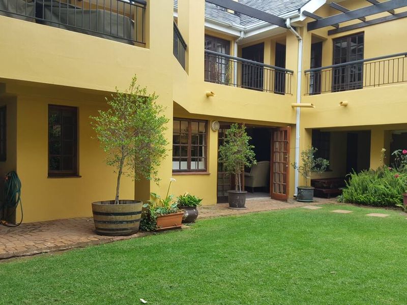 A rear courtyard featuring a manicured lawn, paved patio area, and a two-story building facade with balconies and timber pergola structures.