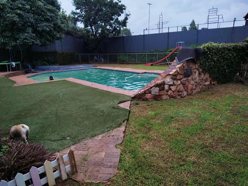 Outdoor pool area featuring a stone feature wall, integrated slide, brick paving, and artificial lawn, enclosed by metal perimeter fencing.