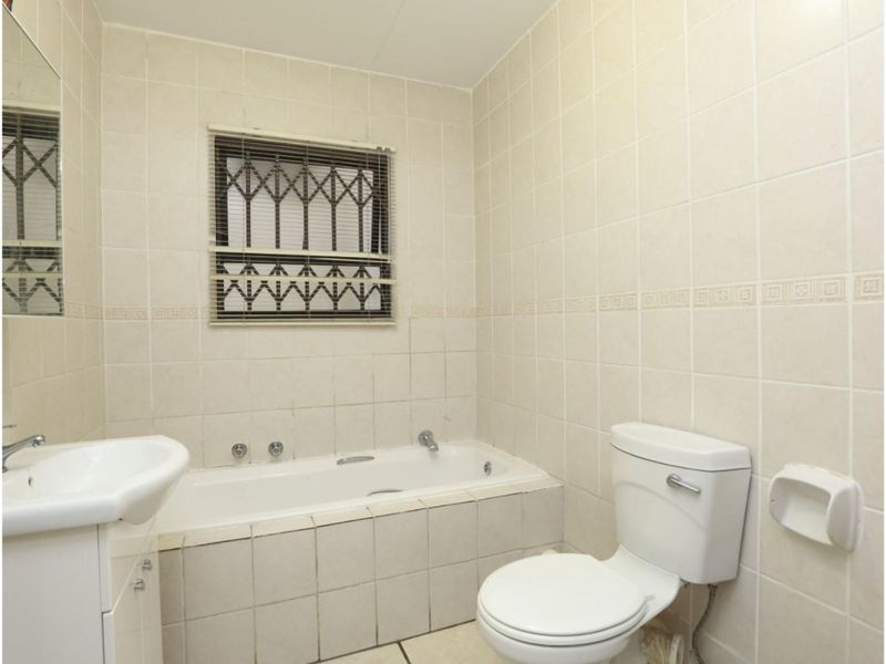 A dated bathroom featuring a built-in bathtub, pedestal basin, and toilet, finished with neutral ceramic wall tiles and a window with security bars.