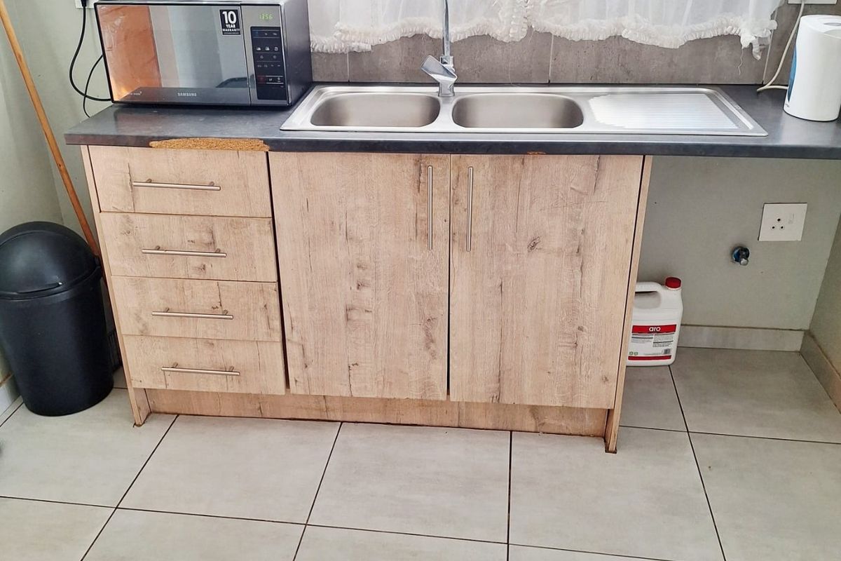 A functional kitchen sink area featuring light wood-look cabinetry, a double stainless steel sink, and tiled flooring, showing signs of wear on the countertop.