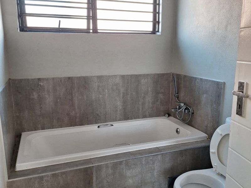 A clean, functional bathroom featuring a built-in bathtub with grey tiled surround, a toilet, and a window with security bars.