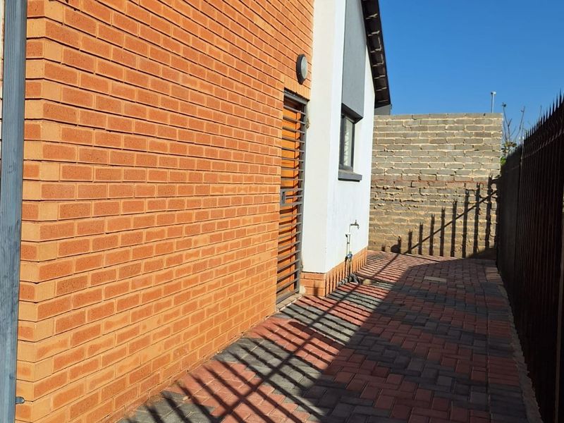 Side passage featuring brick paving, a security gate, and an external water tap, leading to a rear area with a partially finished boundary wall.