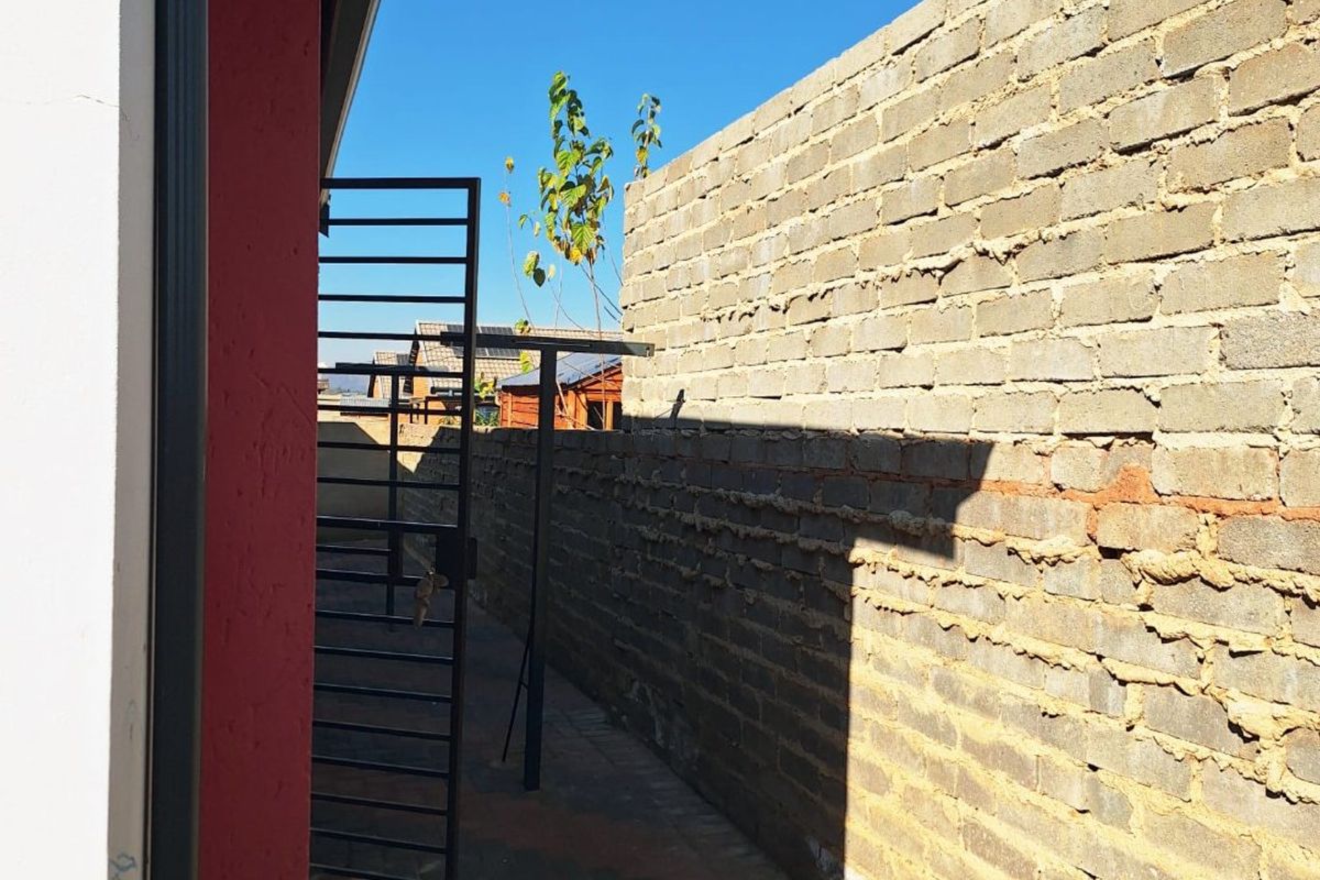 A narrow side passage featuring a paved walkway, metal security gate, and an unfinished brick boundary wall.