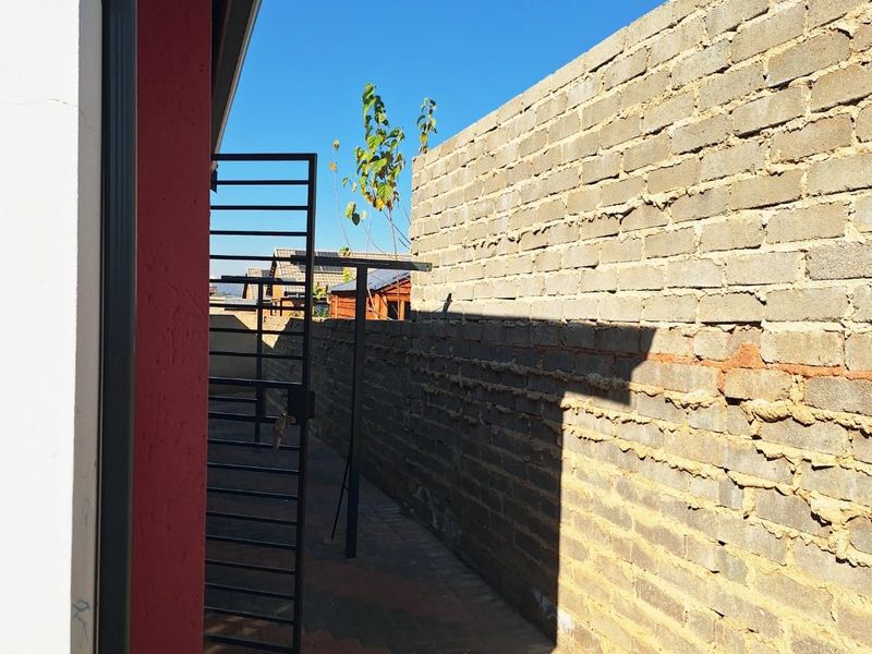 A narrow side passage featuring a paved walkway, metal security gate, and an unfinished brick boundary wall.