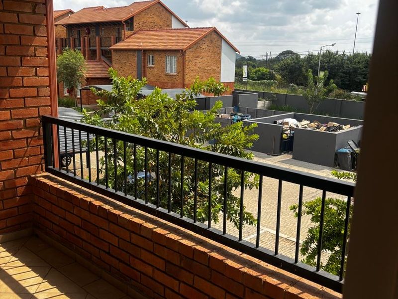 A covered balcony featuring exposed brick walls, tiled flooring, and a metal balustrade, offering an outdoor space with views of the surrounding complex.