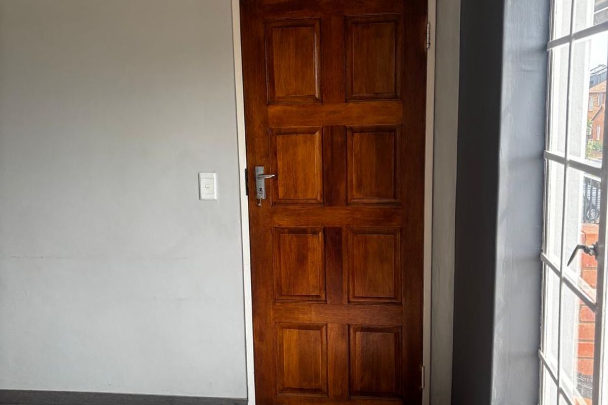 A bright, empty bedroom featuring modern grey laminate flooring, a classic wooden panel door, and a large grid window.