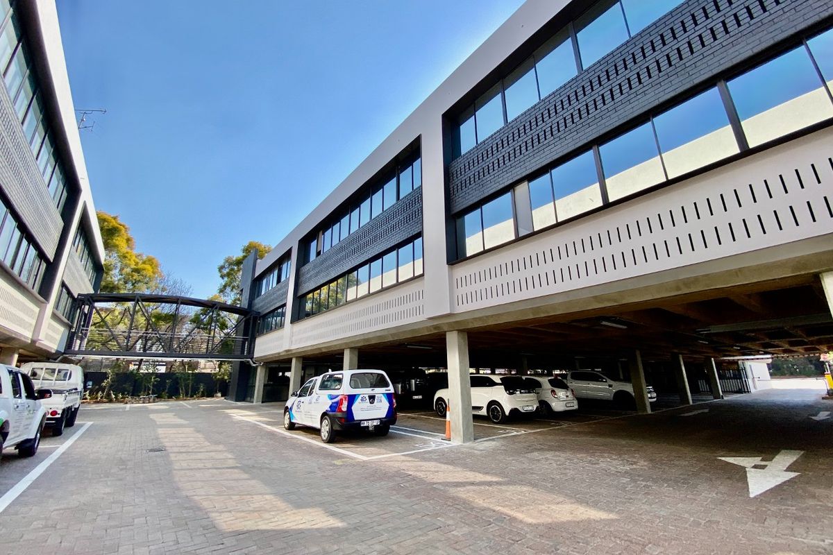 Modern commercial office building featuring a multi-story facade with glass windows, a pedestrian bridge, and a spacious ground-level covered parking area.