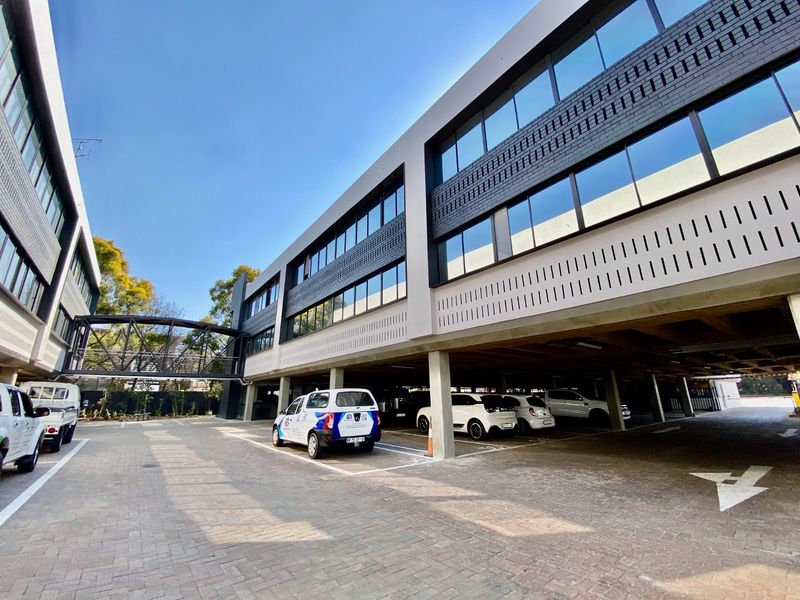 Modern commercial office building featuring a multi-story facade with glass windows, a pedestrian bridge, and a spacious ground-level covered parking area.
