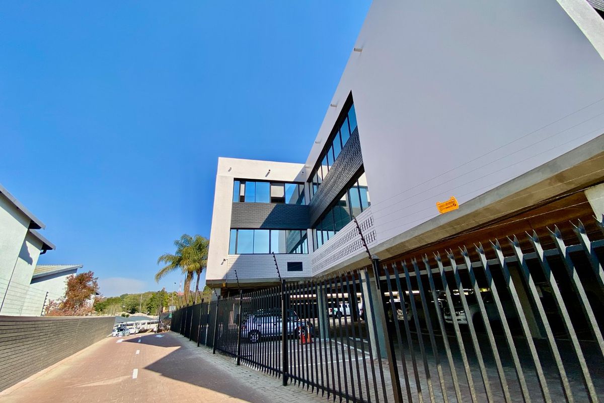 Modern commercial building exterior featuring a paved driveway, secure metal fencing with electric security, and a covered parking area.